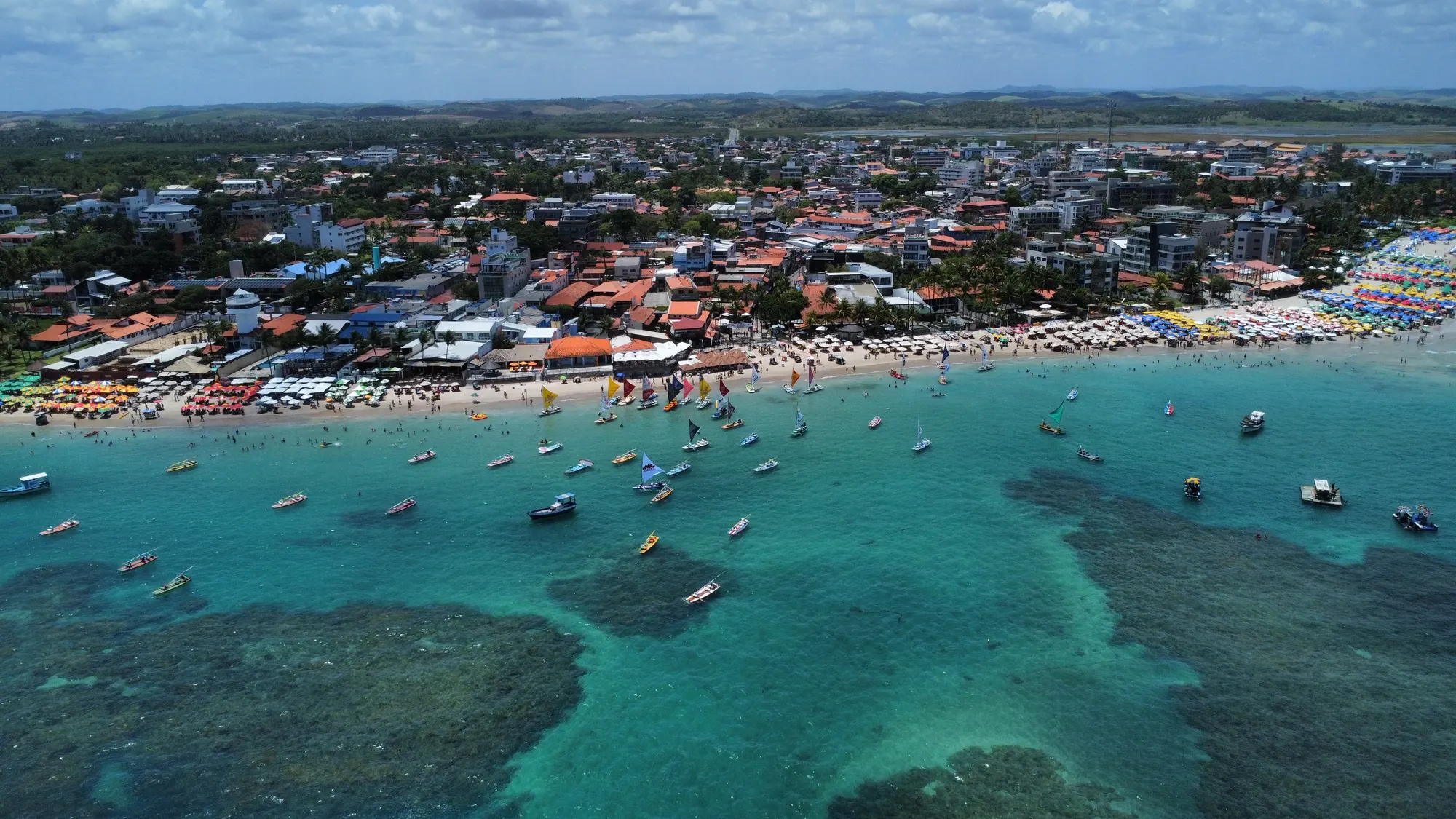 Vista aérea da vila de Porto de Galinhas, com guarda-sóis coloridos na praia, jangadas de velas coloridas e recifes na água turquesa
